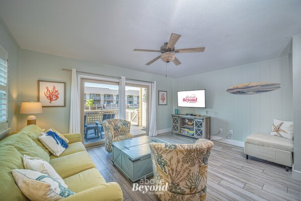 A living room with a green sofa, patterned armchairs, a wooden coffee table, and a ceiling fan. There is a television on a wooden stand and a framed artwork on the wall. - The Cobalt Crab Oasis (Rockport)