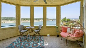 Een kamer met een plafondventilator, een tafel met twee stoelen, een hangstoel met kussens en grote ramen met uitzicht op het strand en de heuvels.