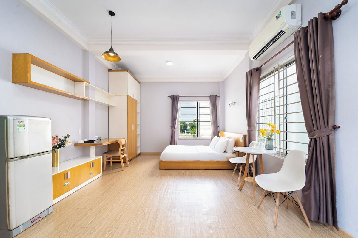 Quarto de hotel compacto com uma cama, uma escrivaninha, uma cadeira, uma mesinha com um vaso de flores e um frigorífico.