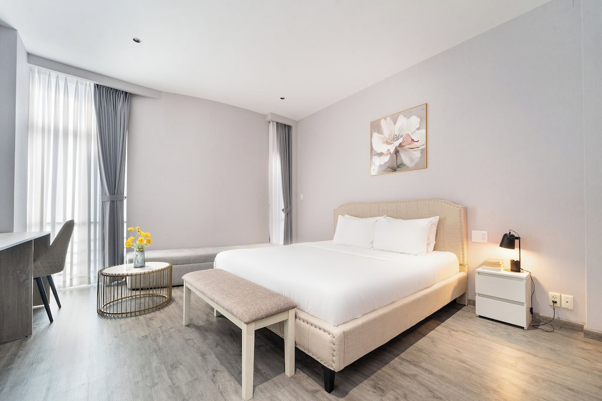 Quarto de hotel moderno com uma cama grande, mesa de cabeceira, banqueta e escrivaninha com cadeira.
