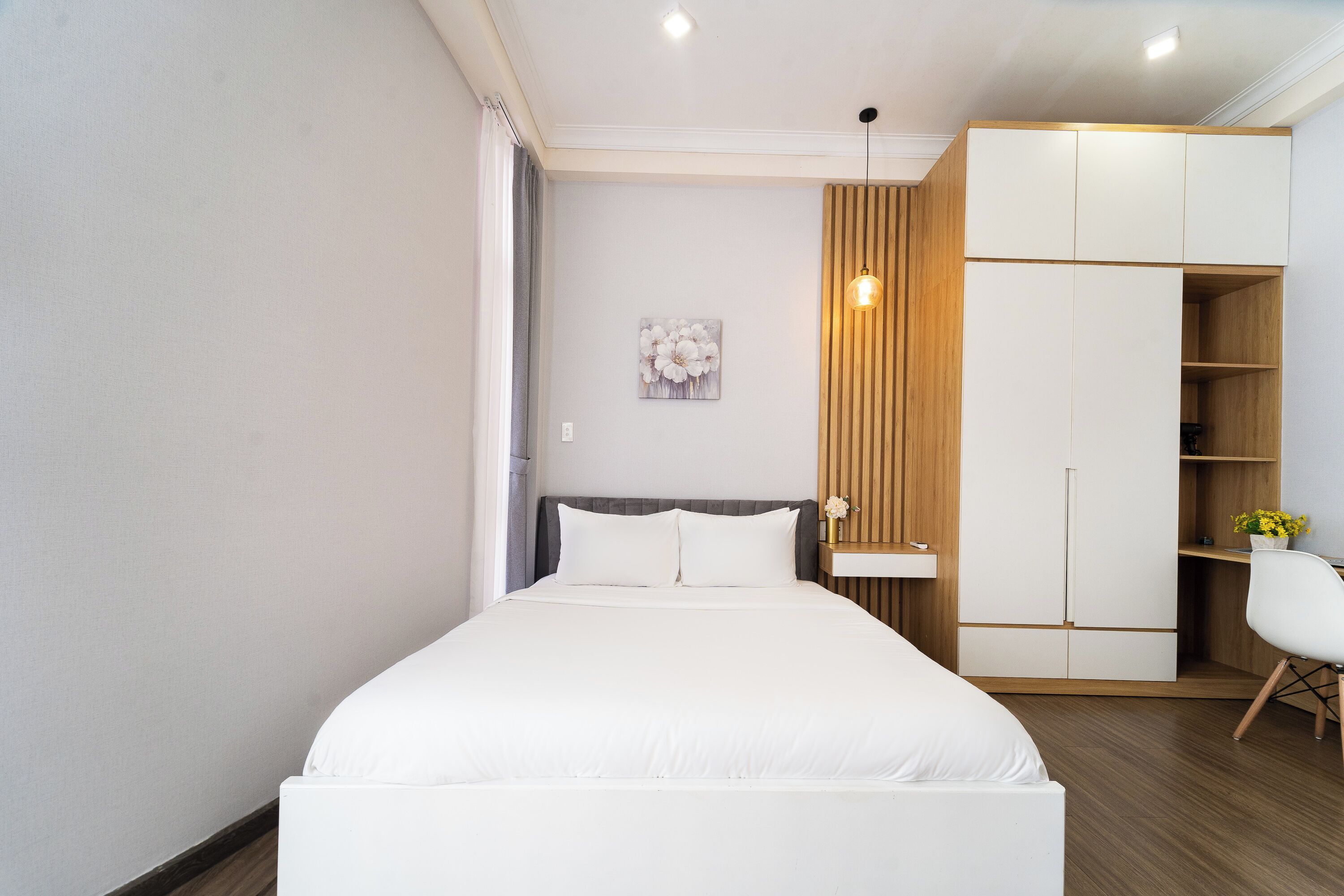 A modern bedroom with a large bed, a wooden wardrobe, a desk, and a hanging light fixture.