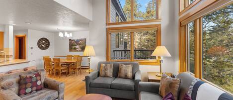 A spacious living area with a grey sofa, a patterned armchair, and a wooden coffee table. There are two lamps on side tables, and a dining area with a wooden table and chairs. Large windows offer a view of trees and a building outside.