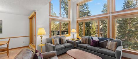 A spacious living room with large windows, a sofa set, a coffee table, and a lamp.
