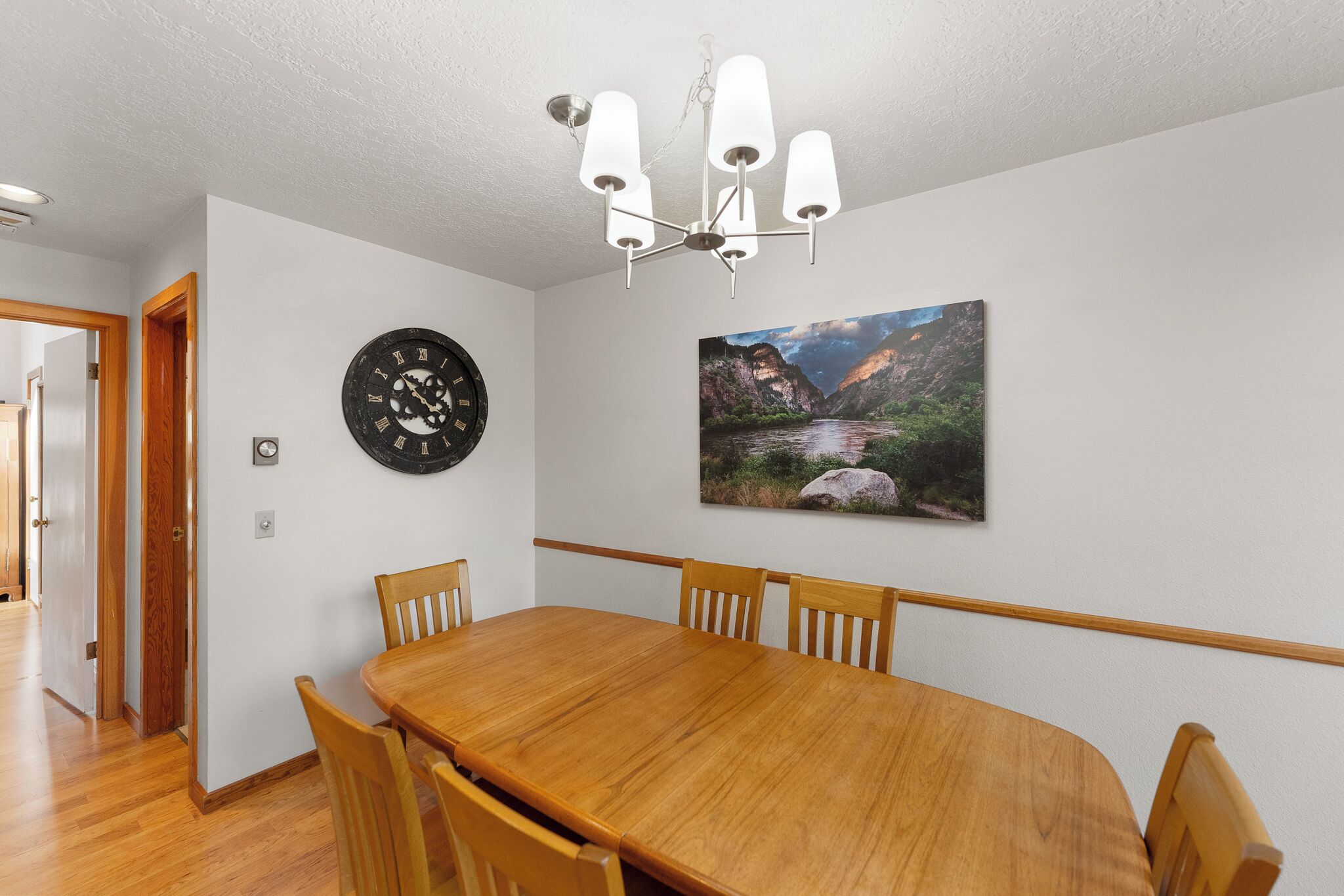 A dining area with a wooden table and chairs, a wall clock, a chandelier, and a landscape painting.