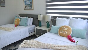 Una habitación de hotel con dos camas, cada una con ropa blanca y almohadas decorativas, una mesita de noche con lámpara y un cuadro en la pared.