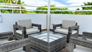 Un patio con mobiliario de mimbre y una mesa con superficie de vidrio, rodeado de paredes blancas y vistas a zonas verdes.