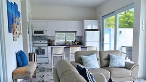 Una cocina moderna con armarios blancos, un microondas y un refrigerador. Hay una zona de comedor con dos sillas blancas y un banco. La habitación tiene un amplio ventanal corredero que da a un espacio exterior.