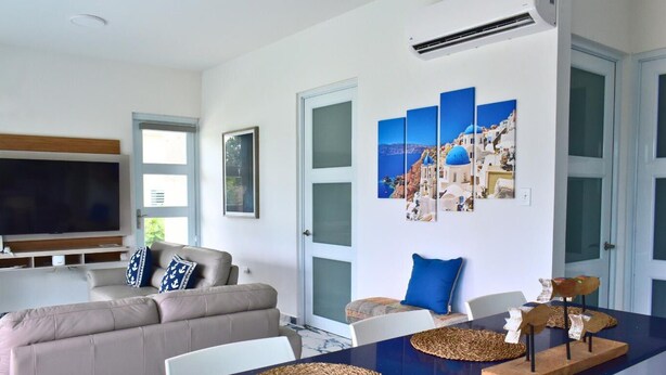Una sala de estar moderna con un televisor de pantalla plana grande, un sofá color beige y un comedor con una mesa azul.
