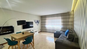 Een woonkamer met een televisie, een bank, een eettafel met stoelen en een groot raam met gordijnen.