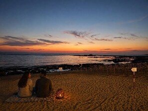 A beach with two people sitting on the sand, a lantern, and a signpost.