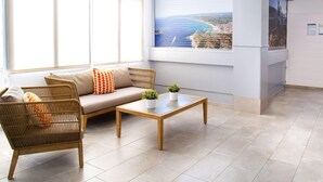 A reception area with a sofa, a wicker chair, a wooden coffee table, and a potted plant. A large window offers a view of the sea and cliffs.