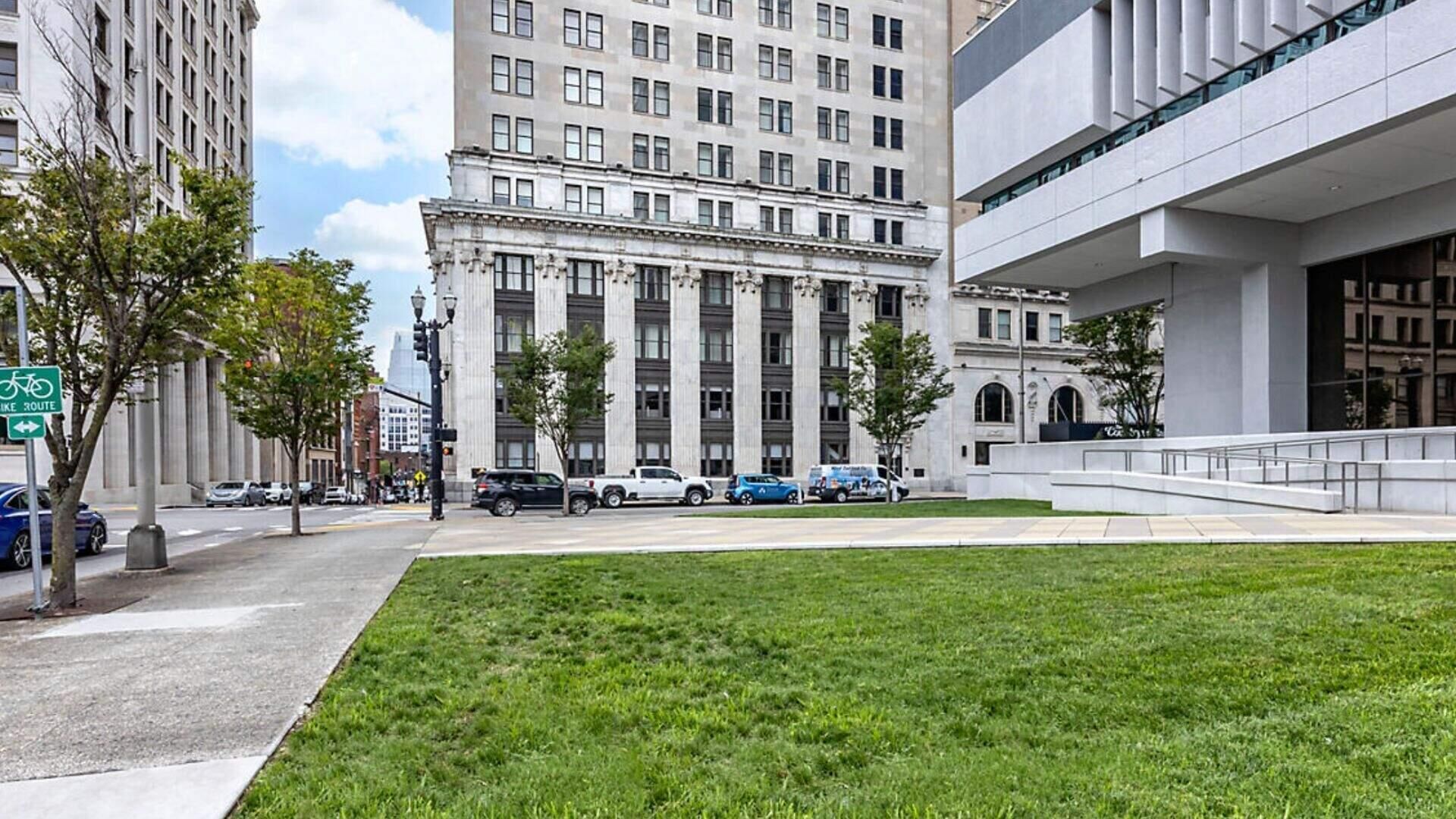 Une zone herbeuse devant un bâtiment à l’architecture classique, un panneau indiquant un parcours cyclable et des voitures garées.