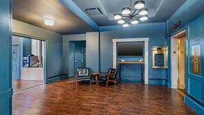 A hotel lobby with a wooden floor, a seating area with chairs, a reception desk, and a blue wall with a mirror.