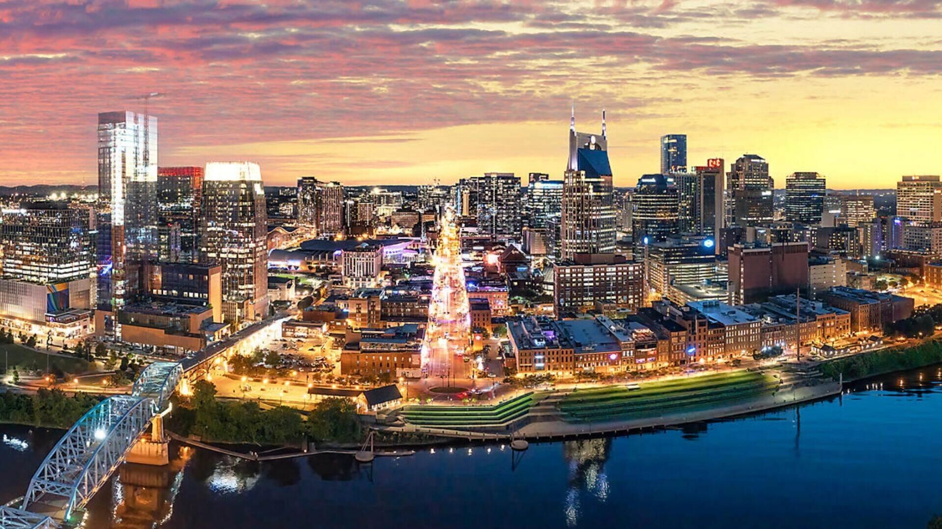 Un paisaje urbano al anochecer con edificios iluminados, un río y un puente.
