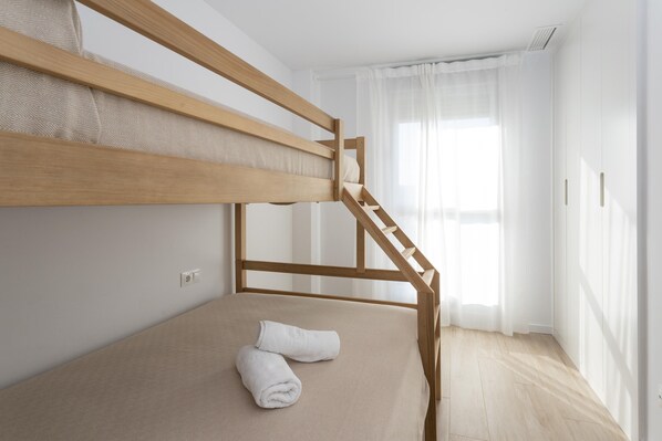 Una cama litera con estructura de madera y ropa de cama color beige, una cortina blanca y un suelo de tonos claros.