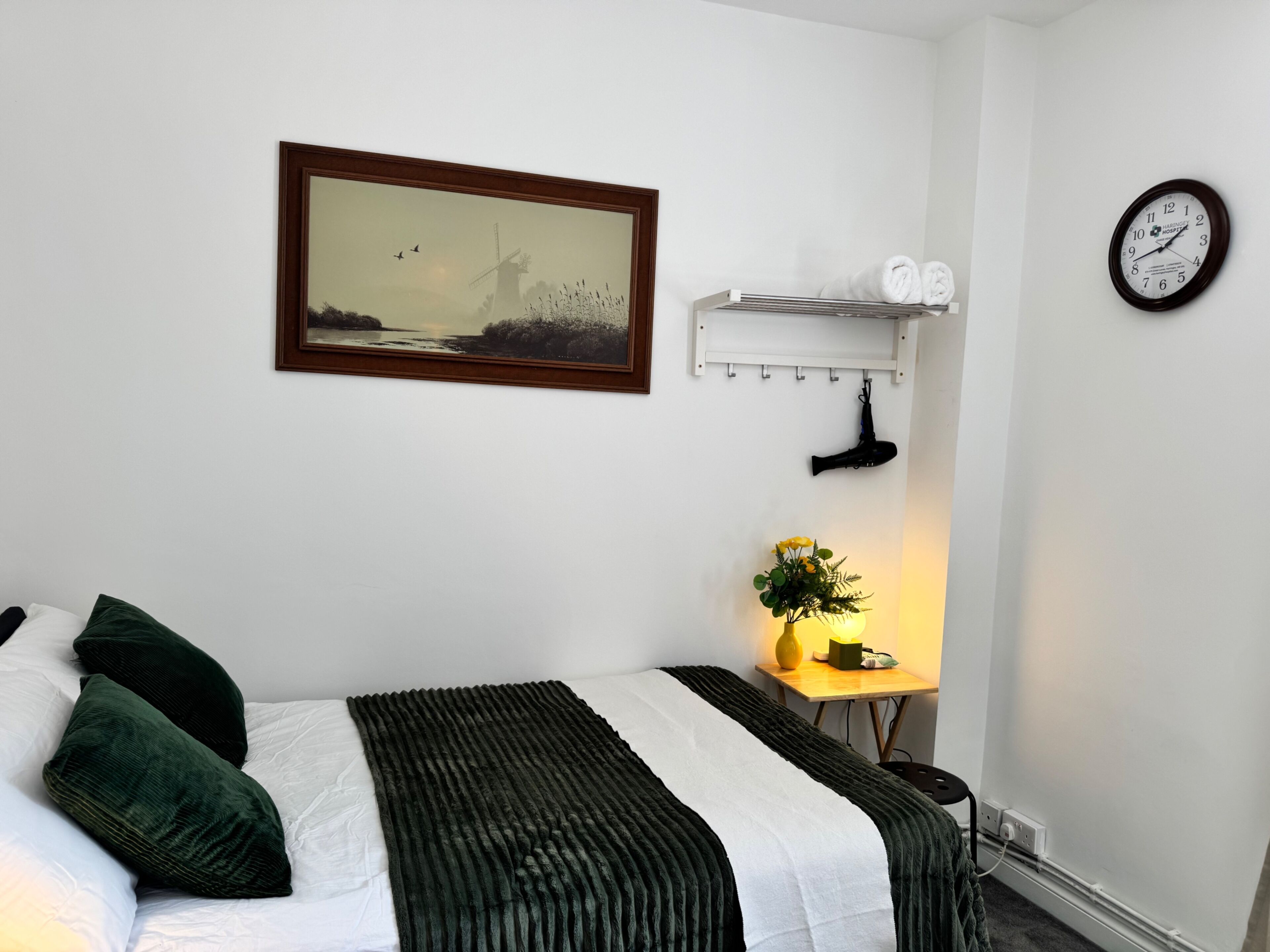 A neatly arranged hotel room with a bed, a wall-mounted clock, a framed picture, and a small table with a vase and a lamp.