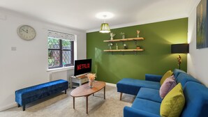 Un salon avec un canapé bleu, une table basse en bois, un banc, un téléviseur affichant le logo « NETFLIX », un mur d’accent vert et une fenêtre donnant sur un paysage.