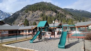 A playground with slides and a fenced area, surrounded by buildings and a mountainous backdrop.