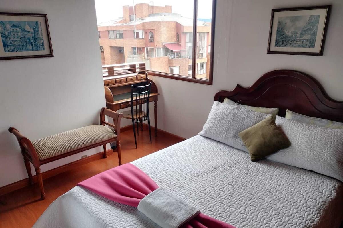 A bedroom with a bed, a desk, a chair, and a window with a view of buildings.