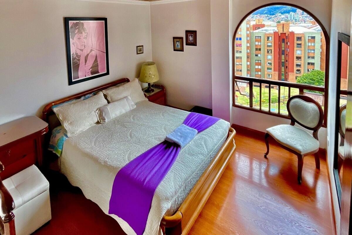 A bedroom with a large bed, a nightstand, a chair, and a window with a city view.