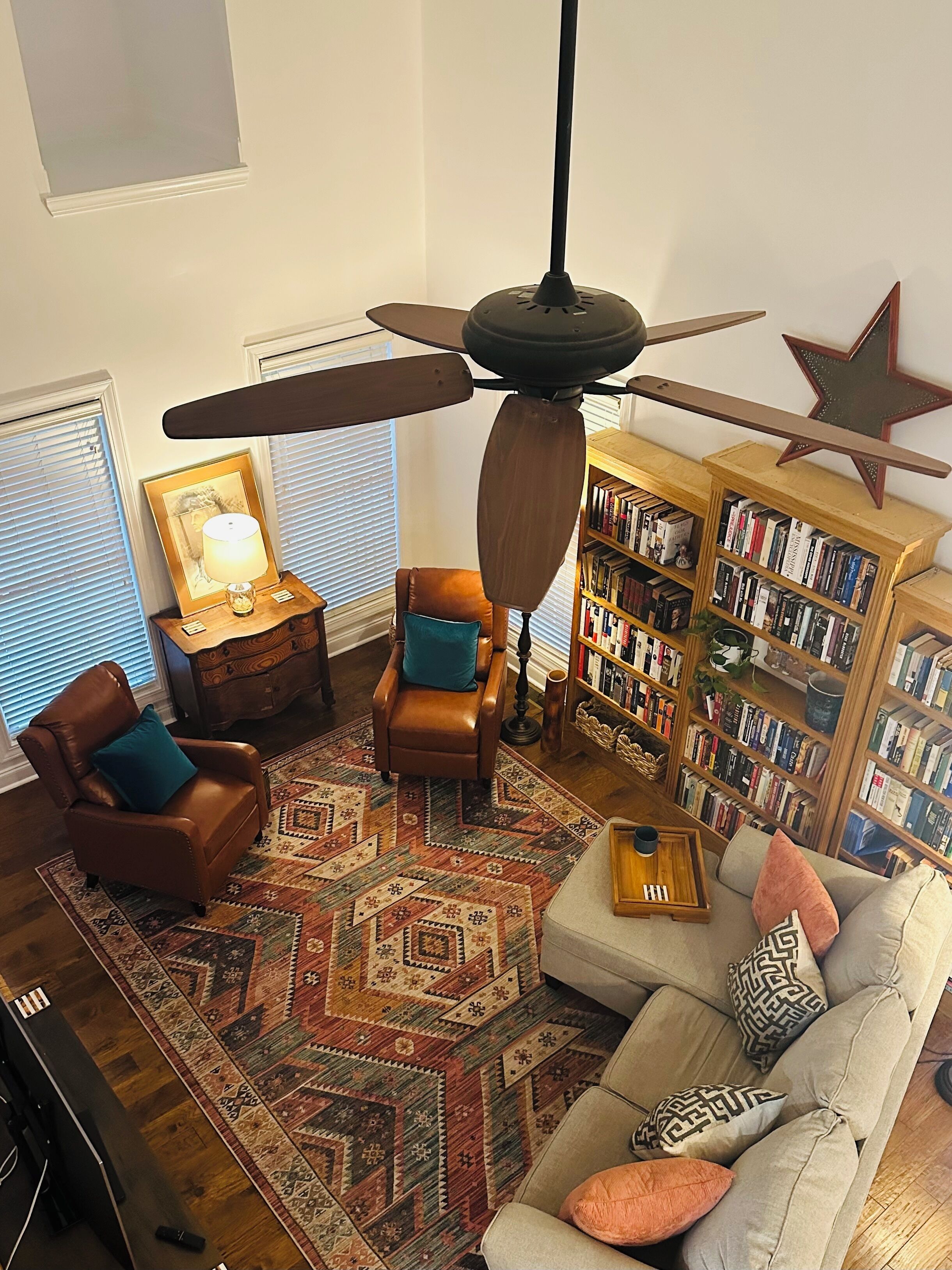 A cozy living room with a ceiling fan, leather armchair, patterned rug, and a bookshelf filled with books.