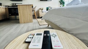 Una habitación de hotel moderna con cama, una mesa redonda de madera con controles remotos, una zona de cocina y un amplio ventanal con vistas al cielo.