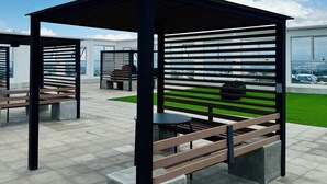 Un espacio de estar al aire libre cubierto, con bancos y mesas, sobre un piso de baldosas y vistas a la ciudad.