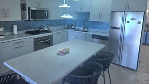 Una cocina moderna con armarios blancos, un refrigerador de acero inoxidable, un microondas y una estufa a gas. Hay dos lámparas colgantes sobre la isla y un lavamanos con grifo.