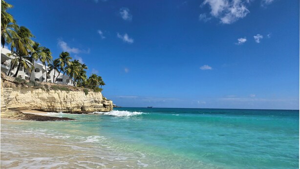Una playa con agua turquesa cristalina, arena blanca y un acantilado con palmeras.