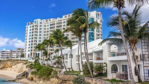 Un hotel frente a la playa con varios balcones, palmeras y un acantilado rocoso.
