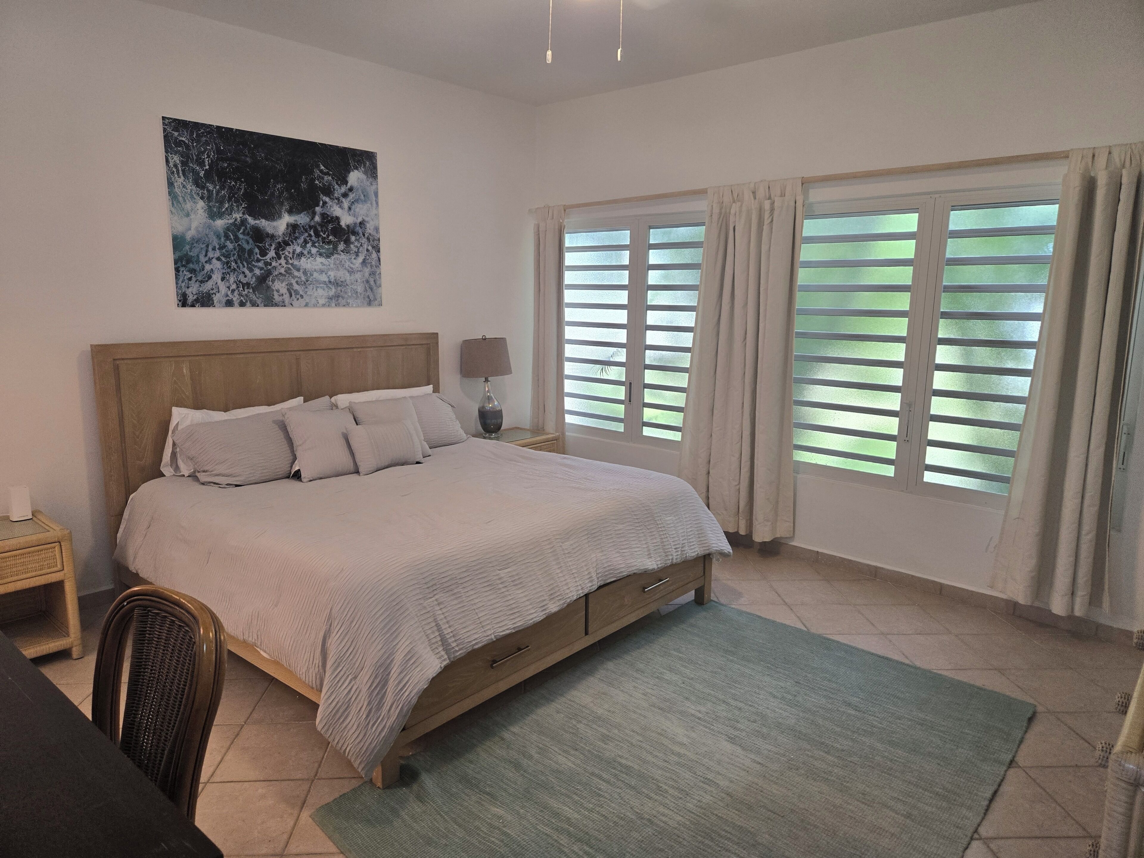 A bedroom with a bed, a nightstand, a lamp, a window with curtains, and a painting on the wall.