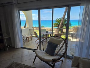 A room with a view of the ocean, a wicker chair, a table, and a small balcony area.