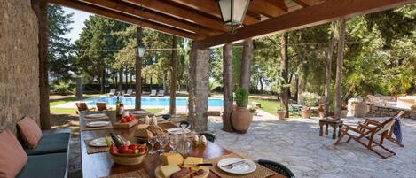 An outdoor dining area with a pool in the background, stone flooring, and wooden furniture.