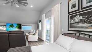 A modern hotel room with a large bed, a sofa, a desk, and a television. There are framed pictures on the wall and a view of the outside through the window.