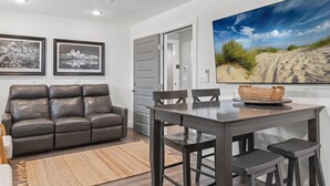 A living room with a dark brown leather sofa, a wooden dining table with two chairs, and a large framed picture of a desert landscape on the wall. - Sugar Sand Cottage B (Port St. Joe)