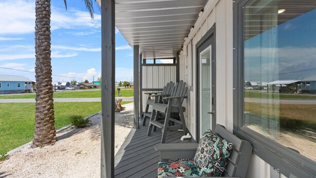 A covered outdoor seating area with a bench and chairs, a palm tree, and a view of a road and buildings. - Sugar Sand Cottage B (Port St. Joe)