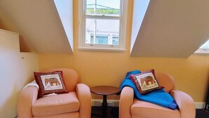 Ein Zimmer mit zwei Sesseln, die ein kleines Tisch abwärts zeigen, einem Fenster mit Blick auf Gebäude und einer geneigten Decke.