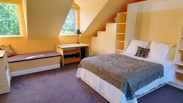 Ein Schlafzimmer mit schrägverdeckter Decke, einem Bett mit gemustertem Überwurf, einem Nachttisch, einem Sessel und einem Fenster mit Blick auf Grünflächen.