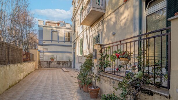 A courtyard with potted plants, a metal fence, and a building with a balcony. - Donna Anna's House by Rentbeat (Civitavecchia)