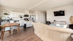 A modern living room with a beige sofa, wooden dining table, and chairs. A kitchen with white cabinets and appliances is visible in the background. - Top floor corner condo with UNREAL views of Commencement Bay + DT Tacoma skyline (Tacoma)