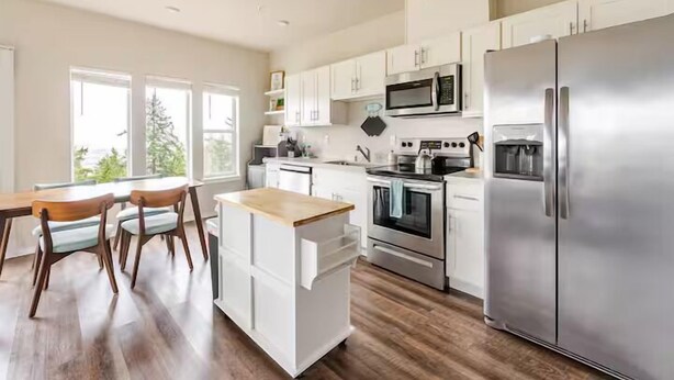 A modern kitchen with white cabinets, stainless steel appliances, and a wooden dining table with chairs. - Top floor corner condo with UNREAL views of Commencement Bay + DT Tacoma skyline (Tacoma)