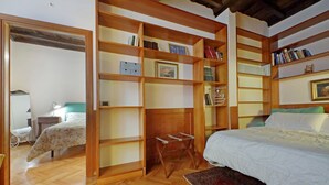 Une chambre avec une bibliothèque en bois, un lit recouvert d’une couette blanche et un petit tabouret en bois.