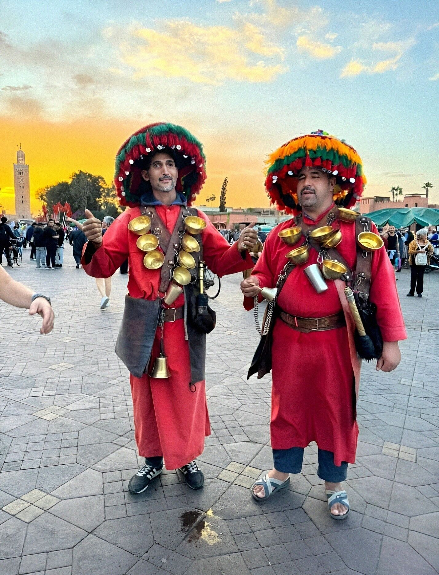 Twee personen in traditionele kleding, met klokken en een toren op de achtergrond.