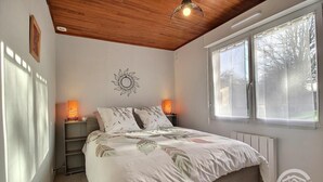 A bedroom with a wooden ceiling, a bed with a floral bedspread, two bedside lamps, a wall-mounted sun decoration, and a window with a view of trees.