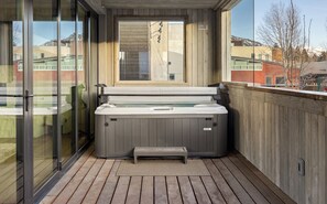 A hot tub on a wooden deck with glass doors and a view of buildings in the background.