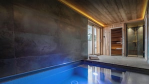 Una piscina interior moderna con agua azul cristalina, techo de madera y puerta de vidrio que da a una terraza de madera.