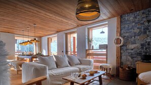 Un salón acogedor y bien iluminado con techo de madera, pared de piedra y ventanas amplias. Hay un sofá, una mesa de centro y una mesa de comedor con sillas.