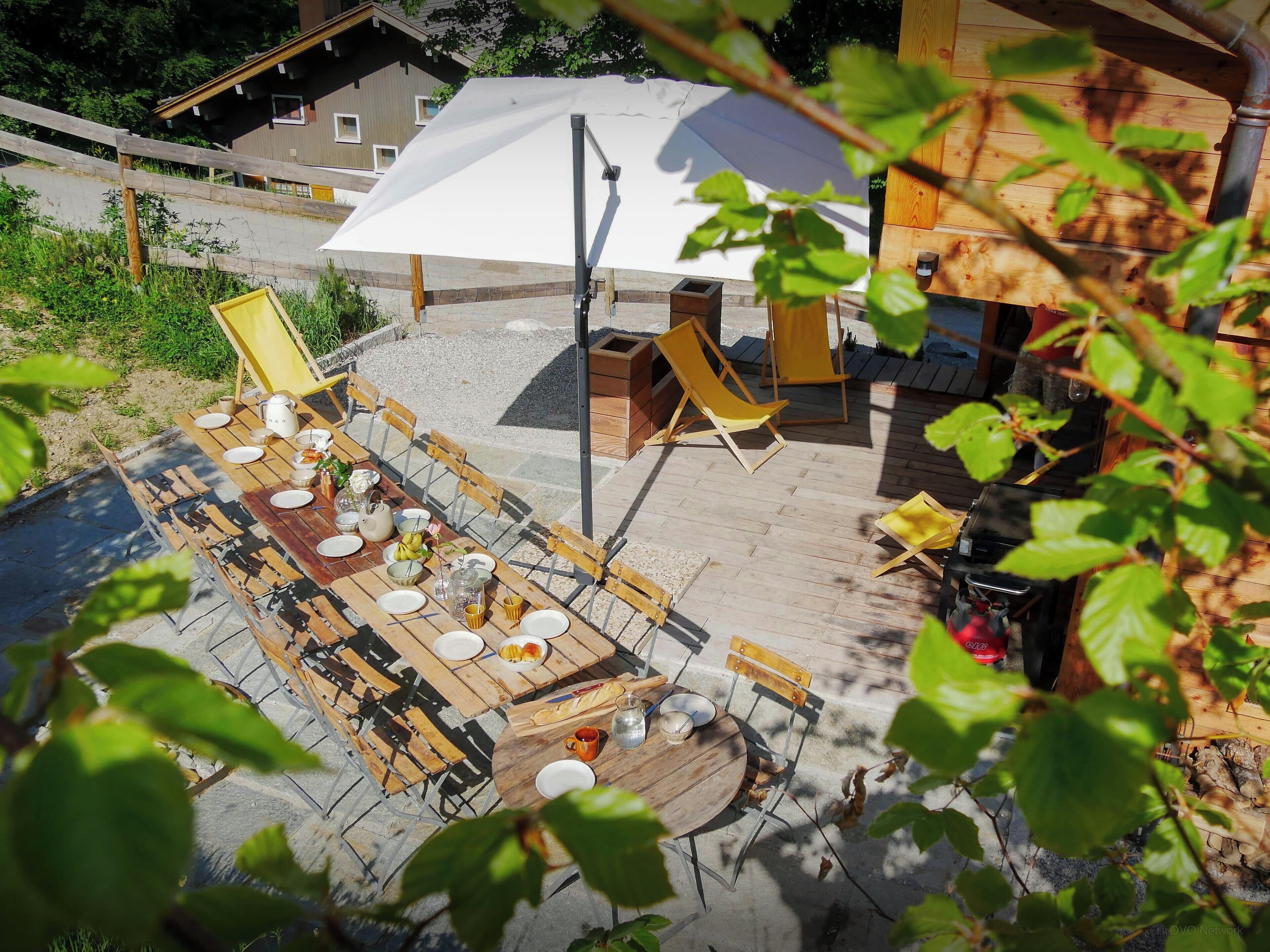 An outdoor dining area with tables and chairs, a white umbrella, and a wooden structure in the background.