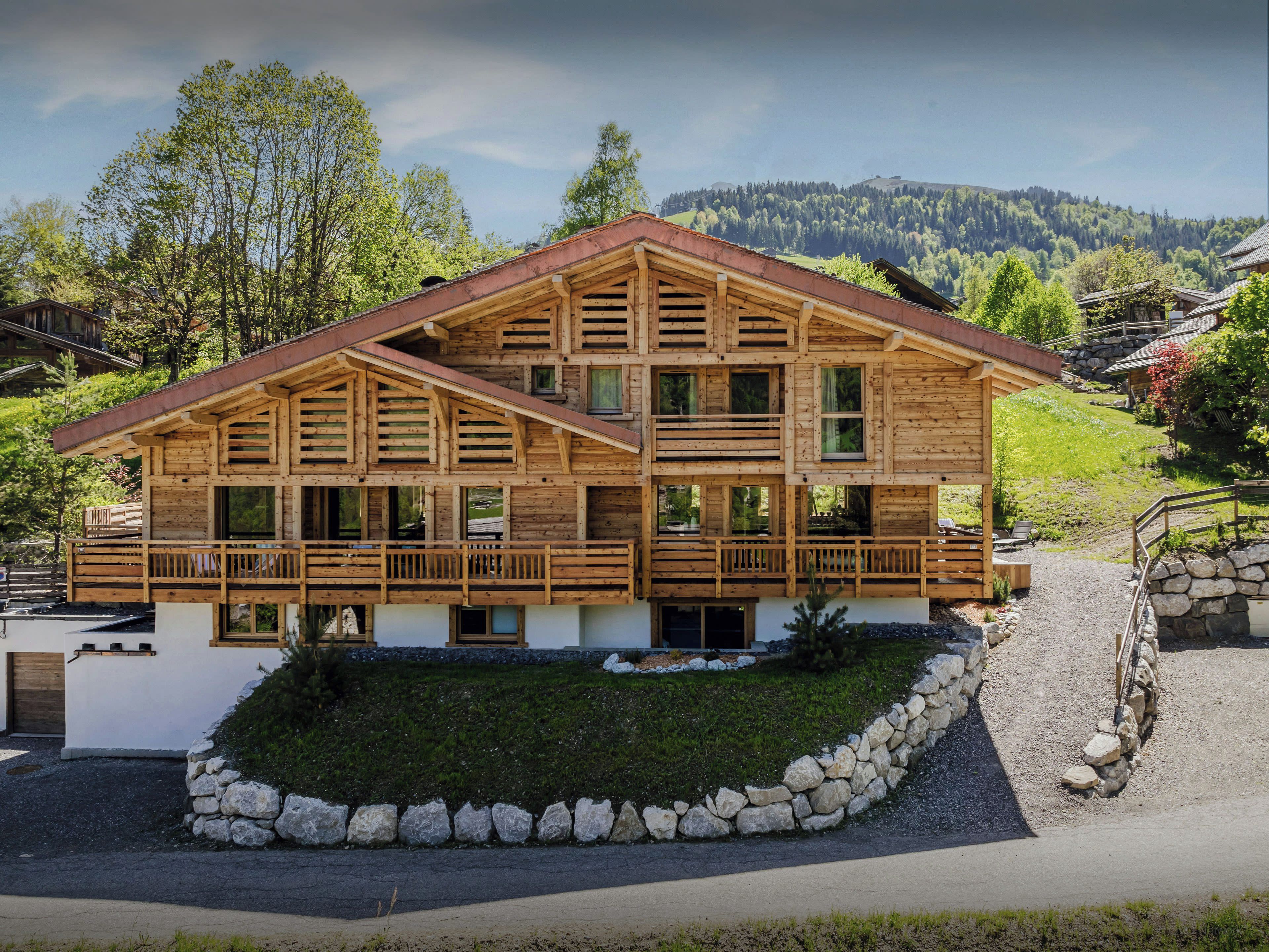 A two-story wooden chalet with a balcony, surrounded by a stone wall and a grassy area.