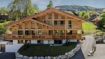 A two-story wooden chalet with a balcony, surrounded by a stone wall and a grassy area.